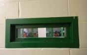 Photo of a small, green viewing window inside a prison. Through the opening are women in their cells wearing orange jumpsuits.