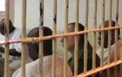 Photo of prison bars and men inside, all facing their backs to the camera.