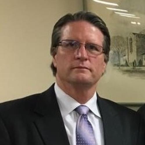 Headshot of Doug Dretke. Short dark hair, glasses, and a dark gray suit.