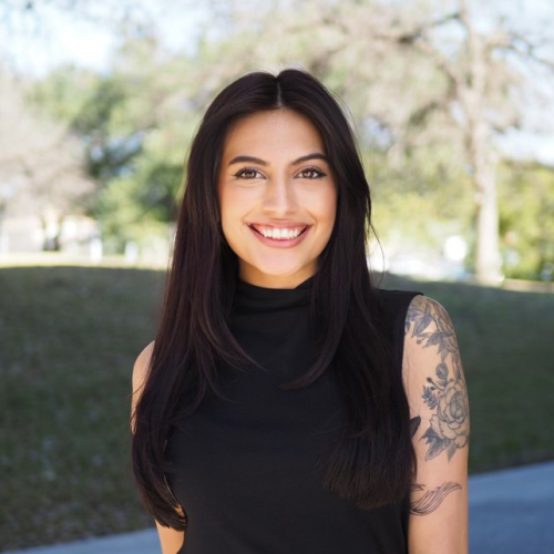 Headshot of Destiny Moreno. Smiling facing the camera, dark long hair, brown eyes, and a tattoo sleeve on her left arm.