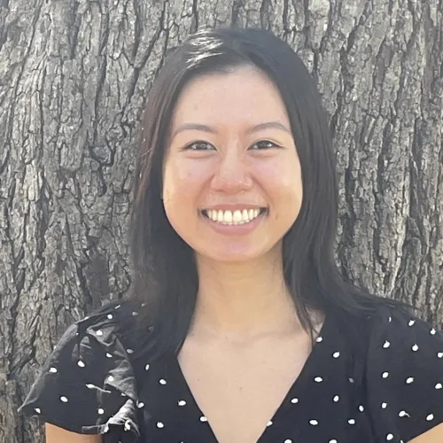 Woman with back hair wearing a black short sleeve top with white dots, smiling facing camera with a tree in the background, headshot for Ashley Nguyen