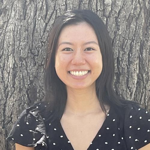 Woman with back hair wearing a black short sleeve top with white dots, smiling facing camera with a tree in the background, headshot for Ashley Nguyen