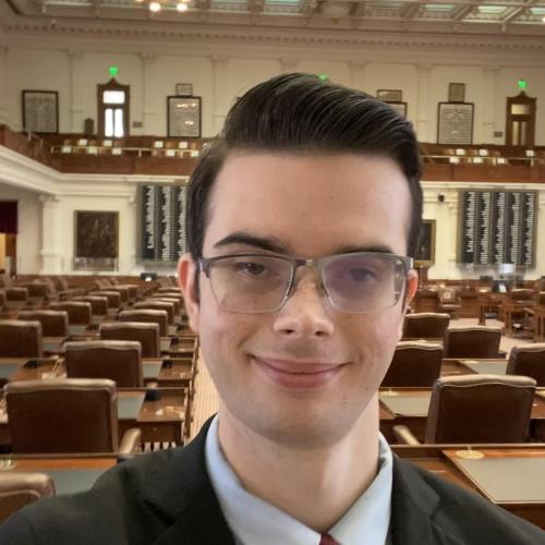 white man with glasses in a black suit, powder blue shirt, and red tie smiling facing the camera with the floor chamber of the Texas House of Representatives in the background