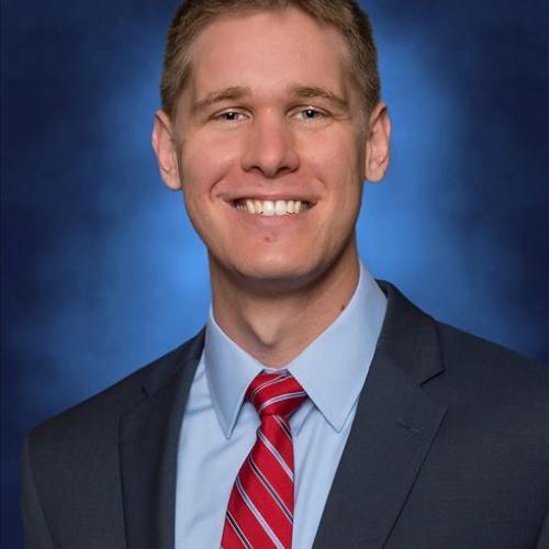 Man smiling facing camera on blue background, in a black suit with a powder blue shirt, and red tie with white and blue stripes, professional headshot of Tyler Sekunda