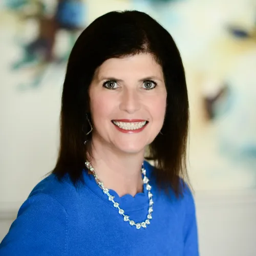 Woman smiling at camera with dark hair and a blue shirt with a pearl necklace on a blurred background